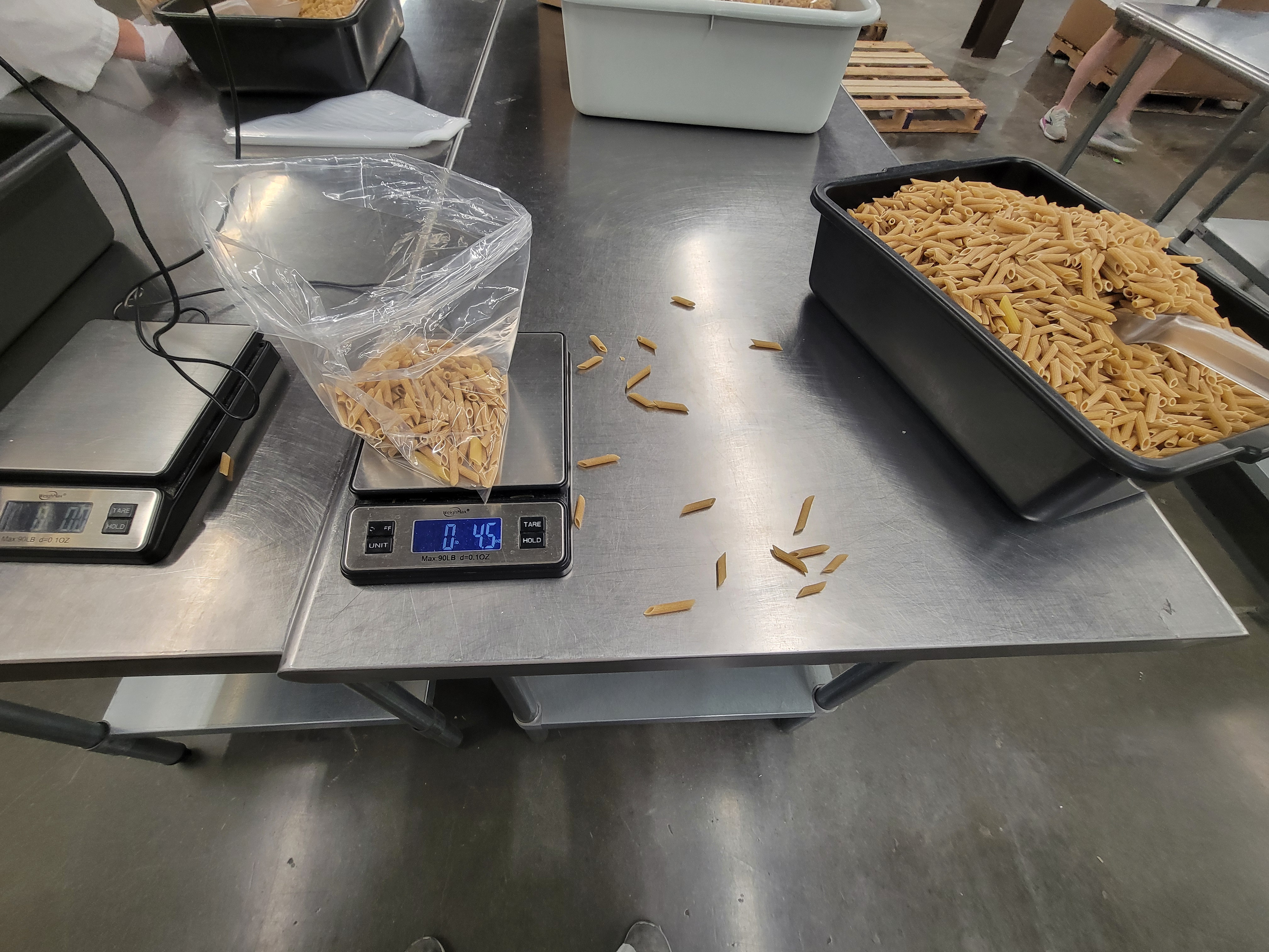 Bag of penne on a scale and tub of penne with some penne spilled between them.